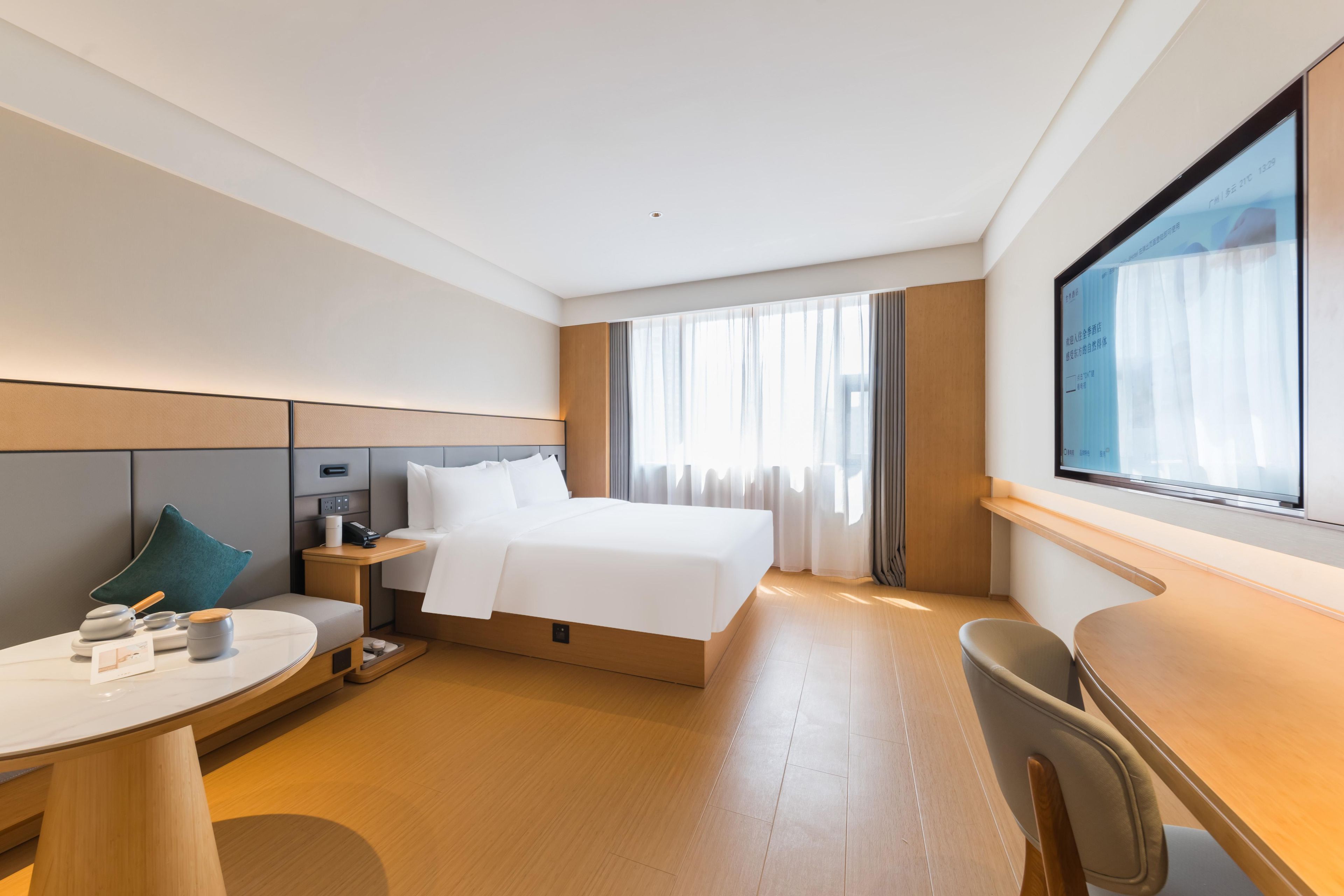 Habitación de hotel moderna con una cama grande, un escritorio y una silla. Hay una mesita de noche con una lámpara y algunos objetos encima. La habitación tiene un ventanal con cortinas.