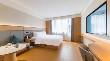 Habitación de hotel moderna con una cama grande, un escritorio y una silla. Hay una mesita de noche con una lámpara y algunos objetos encima. La habitación tiene un ventanal con cortinas.