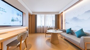 Une chambre d'hôtel moderne, dotée d'un bureau en bois, d'une table ronde, d'un canapé avec des coussins turquoise et d'un grand téléviseur fixé au mur qui affiche un menu.