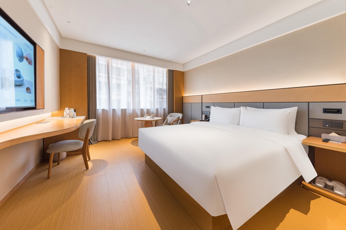 A modern hotel room with a large bed, a desk with a chair, a small round table, and a TV mounted on the wall.