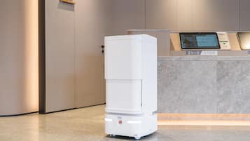 Un quiosco blanco y moderno, de autoservicio, con una pantalla digital, ubicado en un vestíbulo limpio y minimalista con piso de mármol y paredes en tonos neutros.