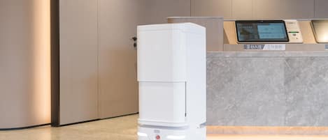 A white, modern, self-service kiosk with a digital display screen, situated in a clean, minimalistic lobby area with a marble floor and neutral-toned walls.