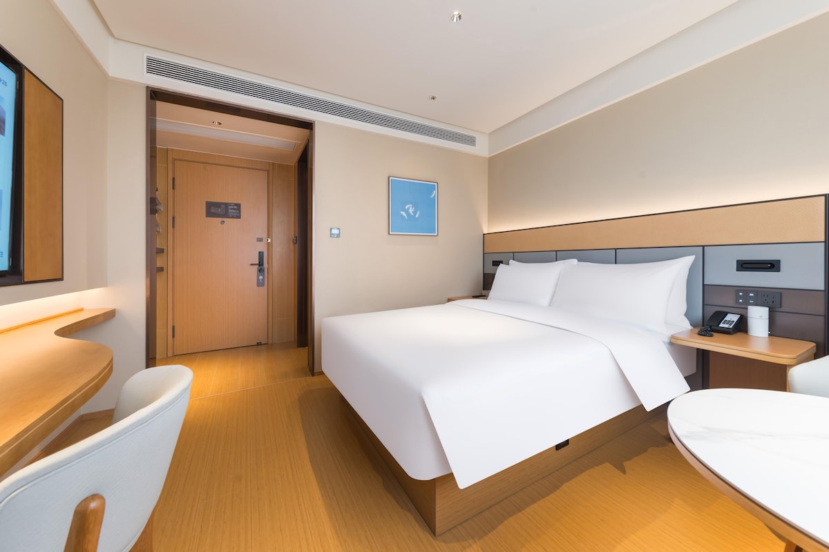 Una habitación de hotel con cama, mesita de noche, silla y puerta que da a otra estancia.
