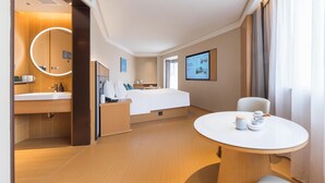 Une chambre d’hôtel moderne avec un plancher en bois, un miroir rond, un lit avec du linge de maison blanc, une table de chevet avec une lampe, une petite table ronde avec des tasses et un téléviseur fixé au mur.