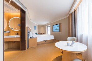A modern hotel room with a wooden floor, a round mirror, a bed with white linens, a bedside table with a lamp, a small round table with cups, and a TV on the wall. - Ji Hotel Guangzhou Shayuan Metro Station (Guangzhou)