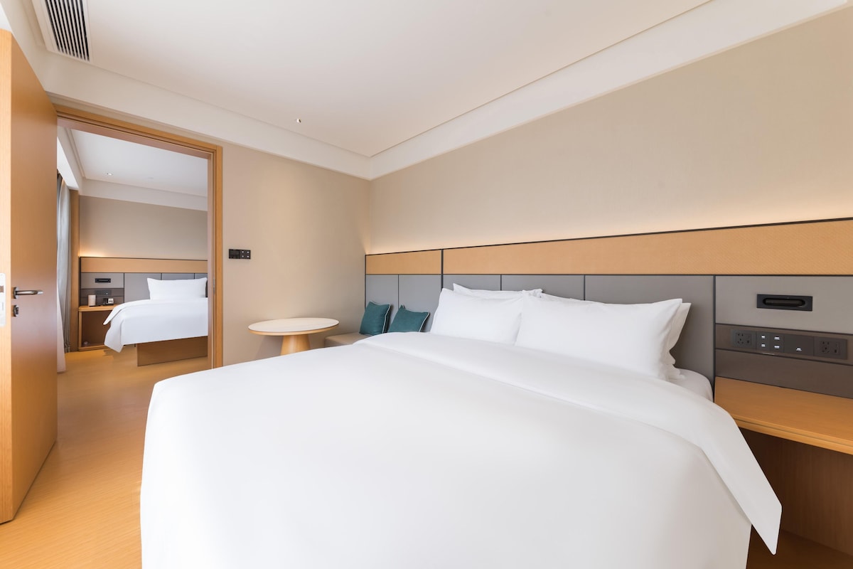 A modern hotel room with a large bed, bedside tables, a round mirror, and a wooden headboard.