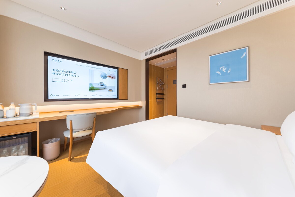 A hotel room with a bed, a desk, a chair, a TV screen, and a framed picture on the wall.
