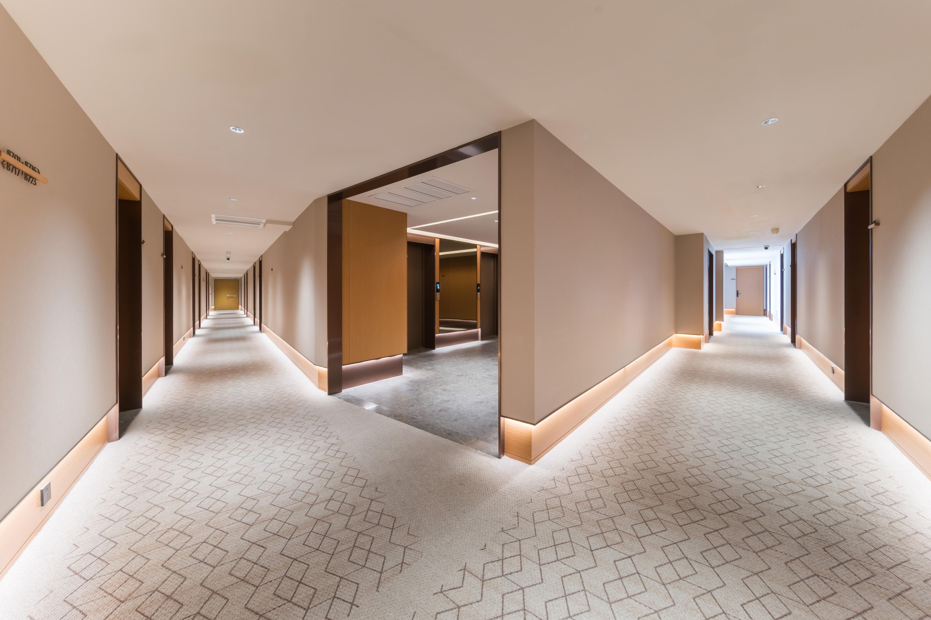 A long, narrow hotel corridor with a patterned carpet, recessed lighting, and a mirrored wall reflecting the corridor.