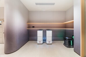 A modern, minimalist bathroom with two white, standalone water dispensers, a black trash bin with a green light, and a wooden shelf above the dispensers. - Ji Hotel Guangzhou Jiangnanxi Metro Station (Guangzhou)