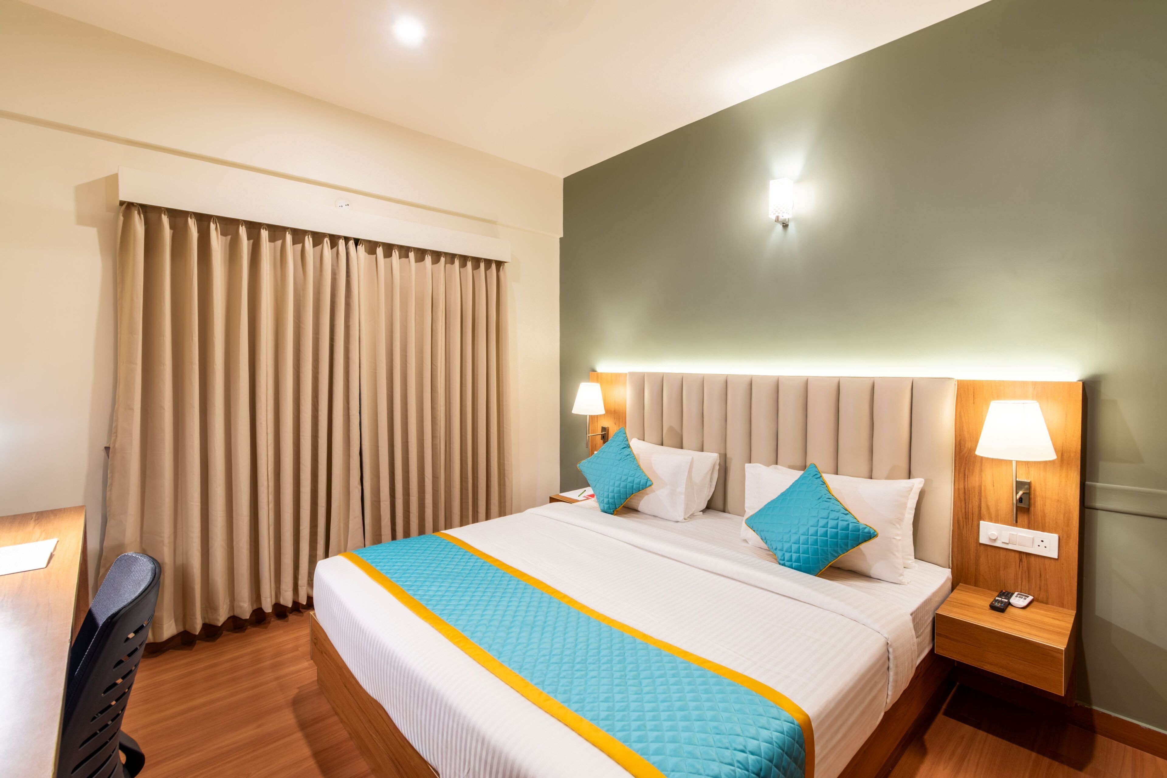 A hotel room with a large bed, two bedside tables with lamps, a desk with a chair, and a window with curtains.