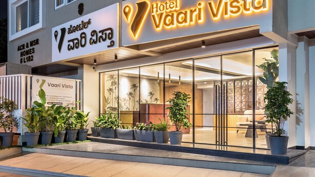 Hotel Vaari Vista con una fachada moderna, un amplio acceso de vidrio y plantas en macetas al frente.