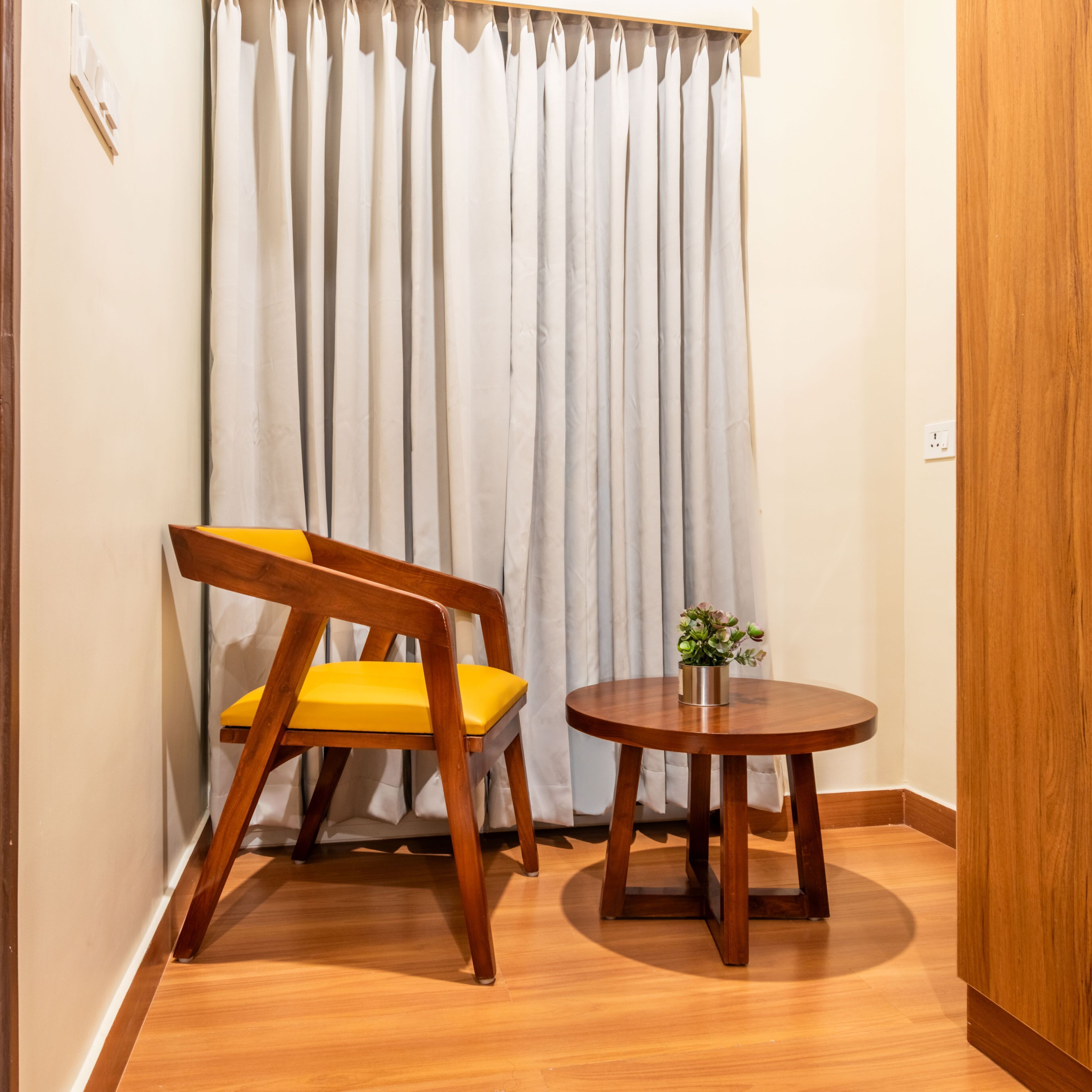 Una habitación pequeña con piso de madera, una silla de madera con cojines amarillos, una mesita redonda con una planta y una cortina blanca.