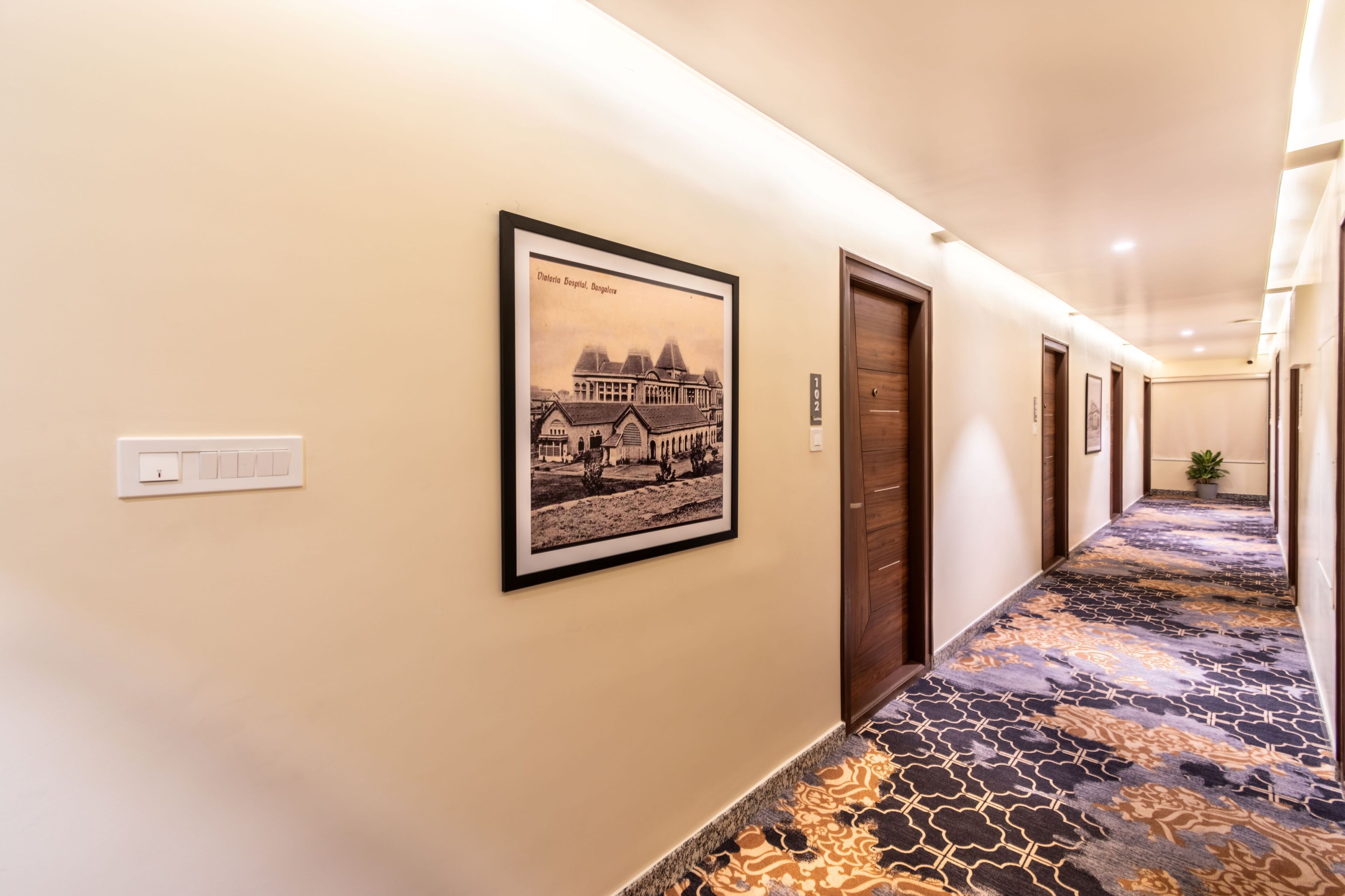 A hotel hallway with a patterned carpet, framed pictures on the wall, and a door on the right.