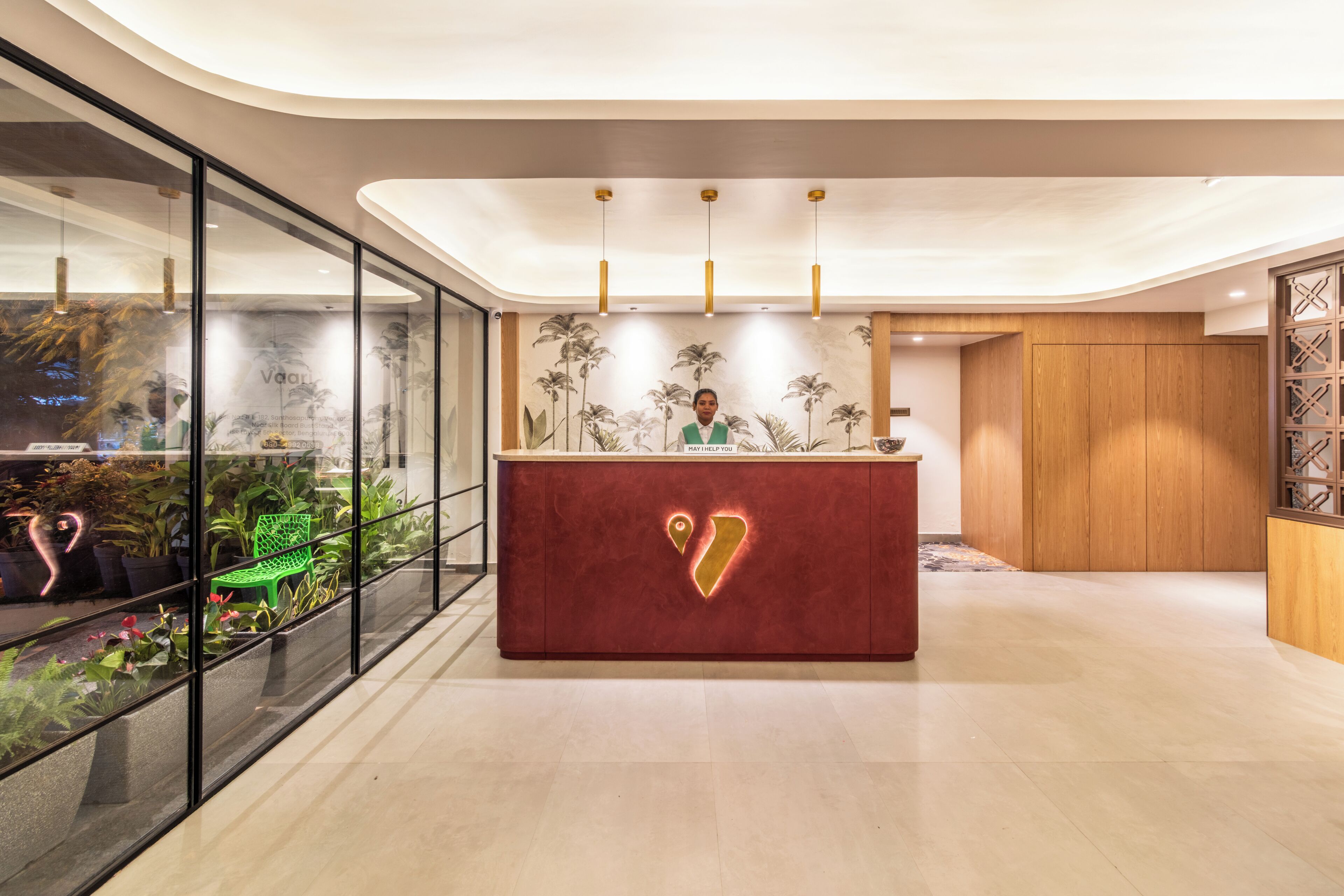 Un área de recepción moderna con piso de mármol, una mesa de recepción roja con logotipo dorado y un muro de vidrio con plantas en macetas.