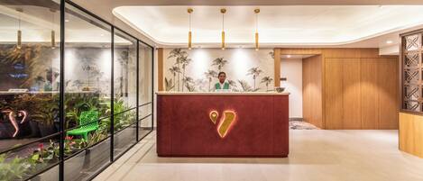 Un área de recepción moderna con piso de mármol, una mesa de recepción roja con logotipo dorado y un muro de vidrio con plantas en macetas.