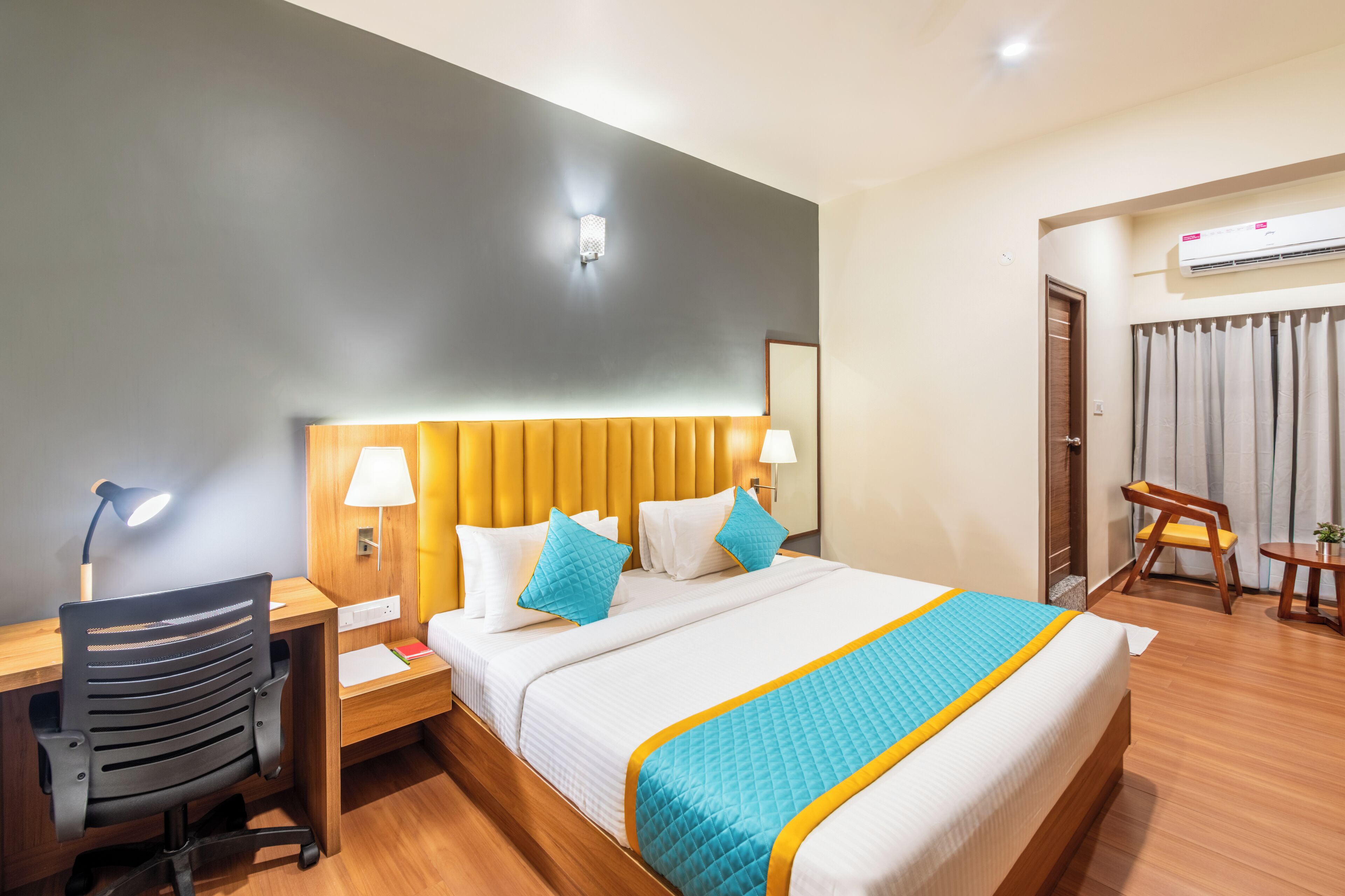 A hotel room with a bed, a desk, a chair, a lamp, and an air conditioning unit.