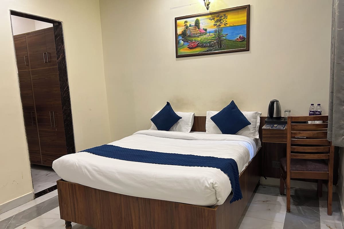 A neatly made bed with a blue and white bedspread, a wooden nightstand with a kettle and water bottles, and a painting of a rural scene on the wall.