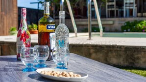 A table with bottles and glasses, a plate with nuts, and a view of a pool and buildings in the background. - Salamandre Guest Room with Shared Pool, Wi-Fi, and Air Conditioning (Port-Saint-Louis-du-Rhône)