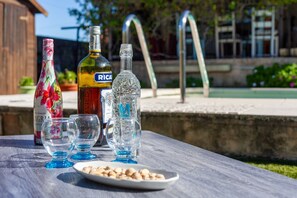 Uma mesa com garrafas e copos, um prato com frutos secos, e uma vista de uma piscina e edifícios ao fundo.