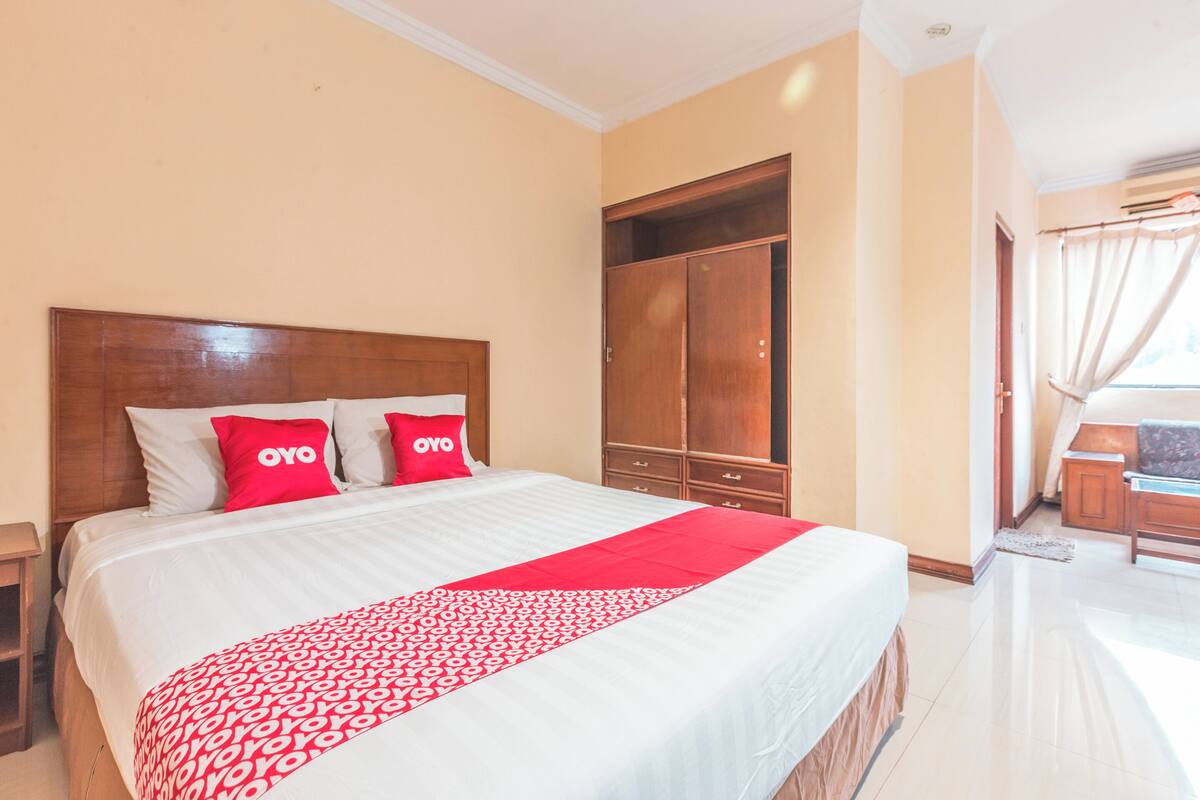 A hotel room with a large bed, two red pillows, a wooden wardrobe, and a view of the outside through a window.