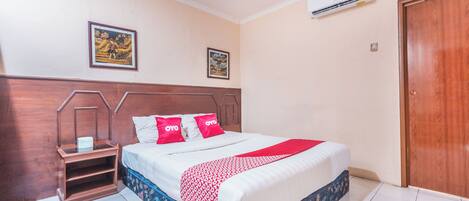 A hotel room with a wooden headboard, a bed with white and red bedding, two framed pictures on the wall, and an air conditioning unit above the bed.