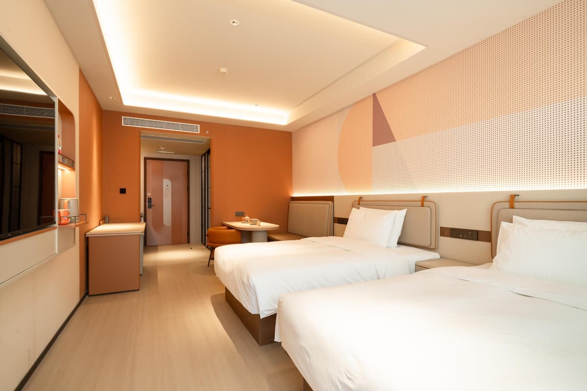 A hotel room with two beds, a small table, a chair, and a large window with a patterned blind.