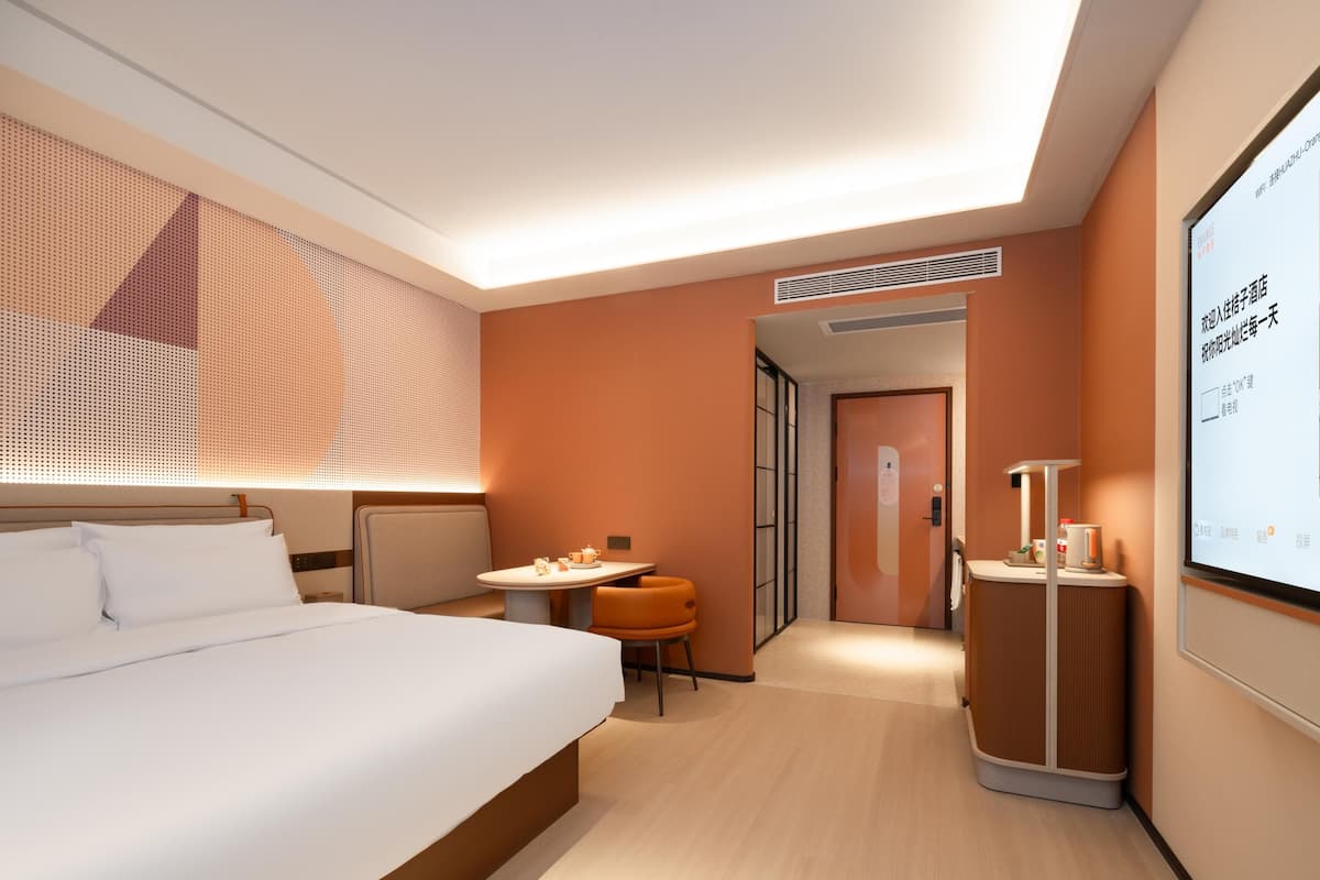 A modern hotel room with a large bed, a small table with a chair, a desk with a chair, and a screen displaying information.