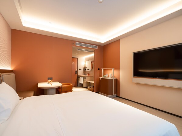 A modern hotel room with a large bed, a TV mounted on the wall, a small round table with a chair, and a bathroom visible through an open door. - Orange Guangzhou Panyu Chimelong Wanbo Center (Guangzhou)