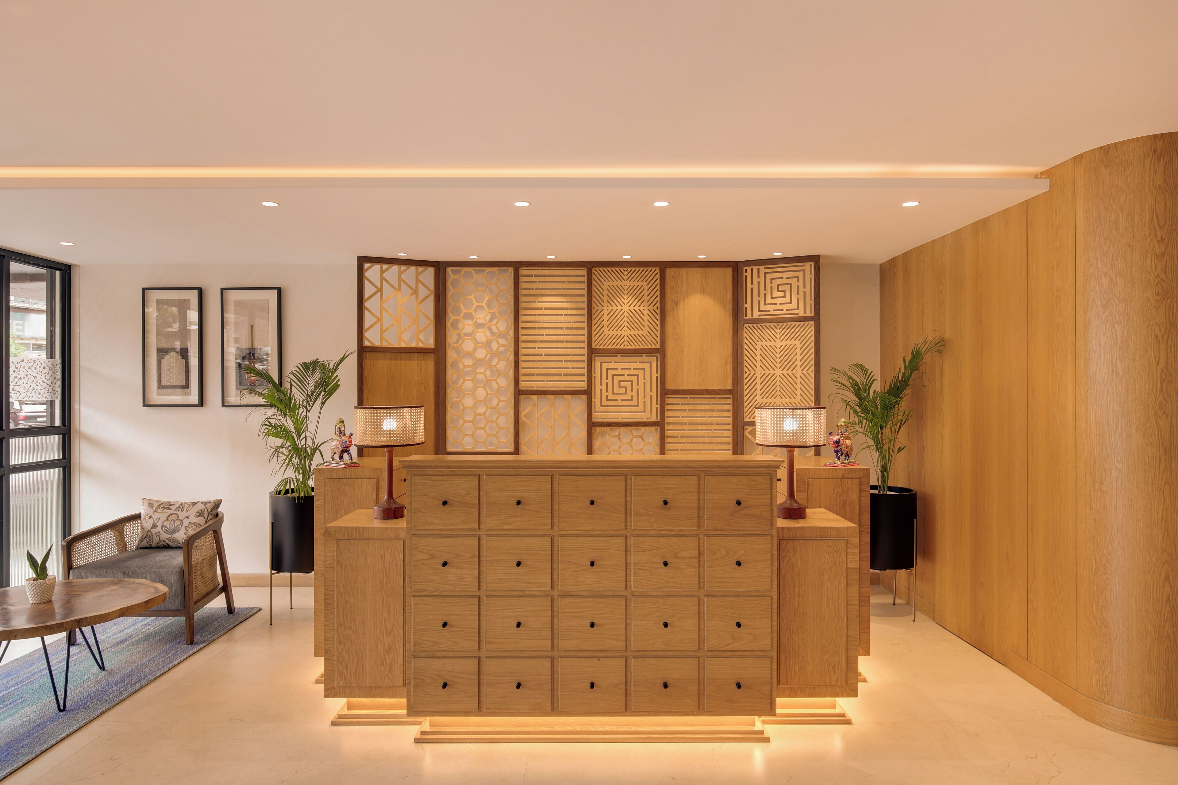 A reception desk with a wooden finish, multiple drawers, and two lamps. There are framed pictures on the wall and a potted plant to the left.