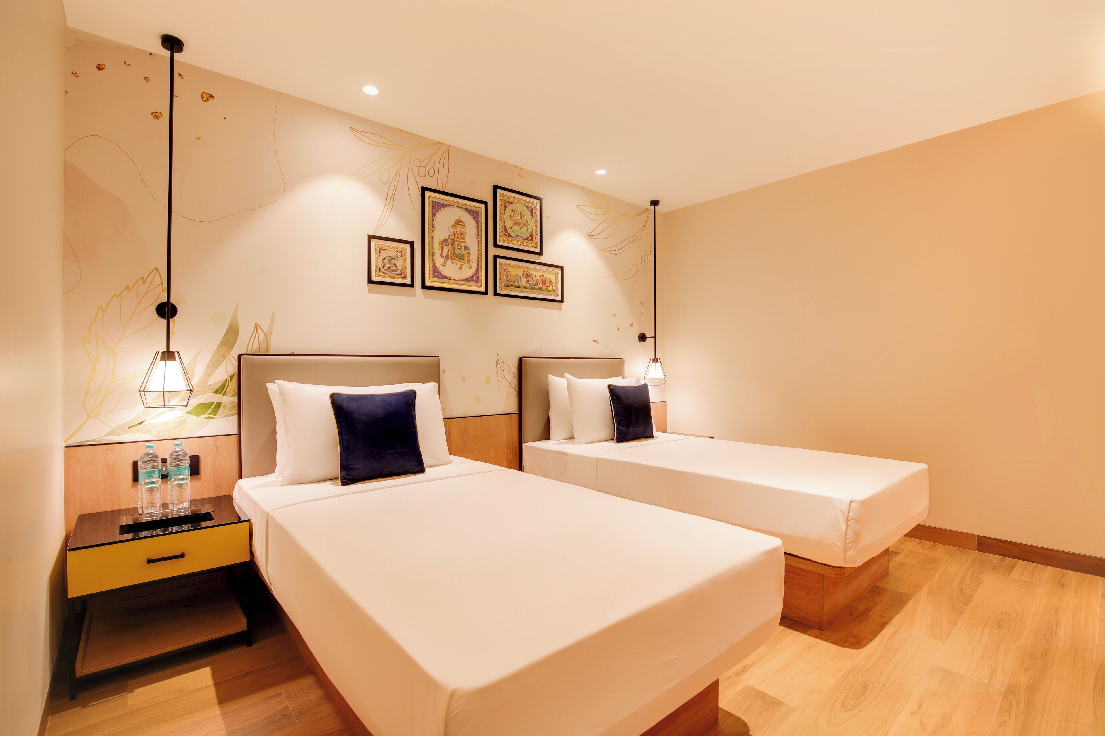 A hotel room with two single beds, a nightstand with a lamp, and a wall with framed pictures.