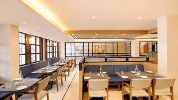 A modern restaurant with long tables, cushioned seating, and large windows.