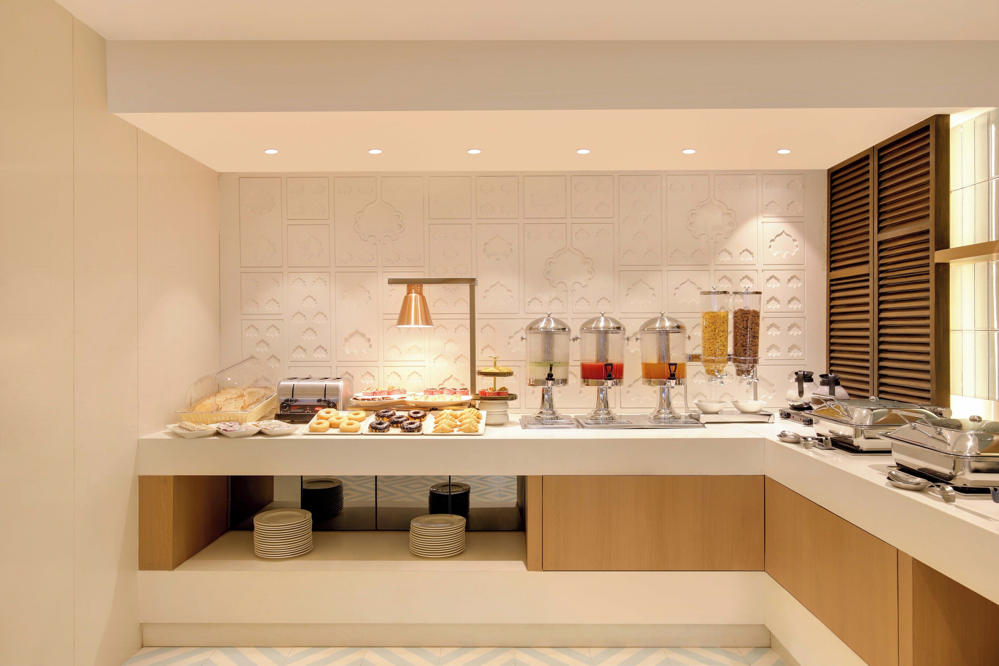 Una barra de desayuno moderna con diversos alimentos y bebidas, una pared decorativa con patrones geométricos y una ventana de listones de madera.