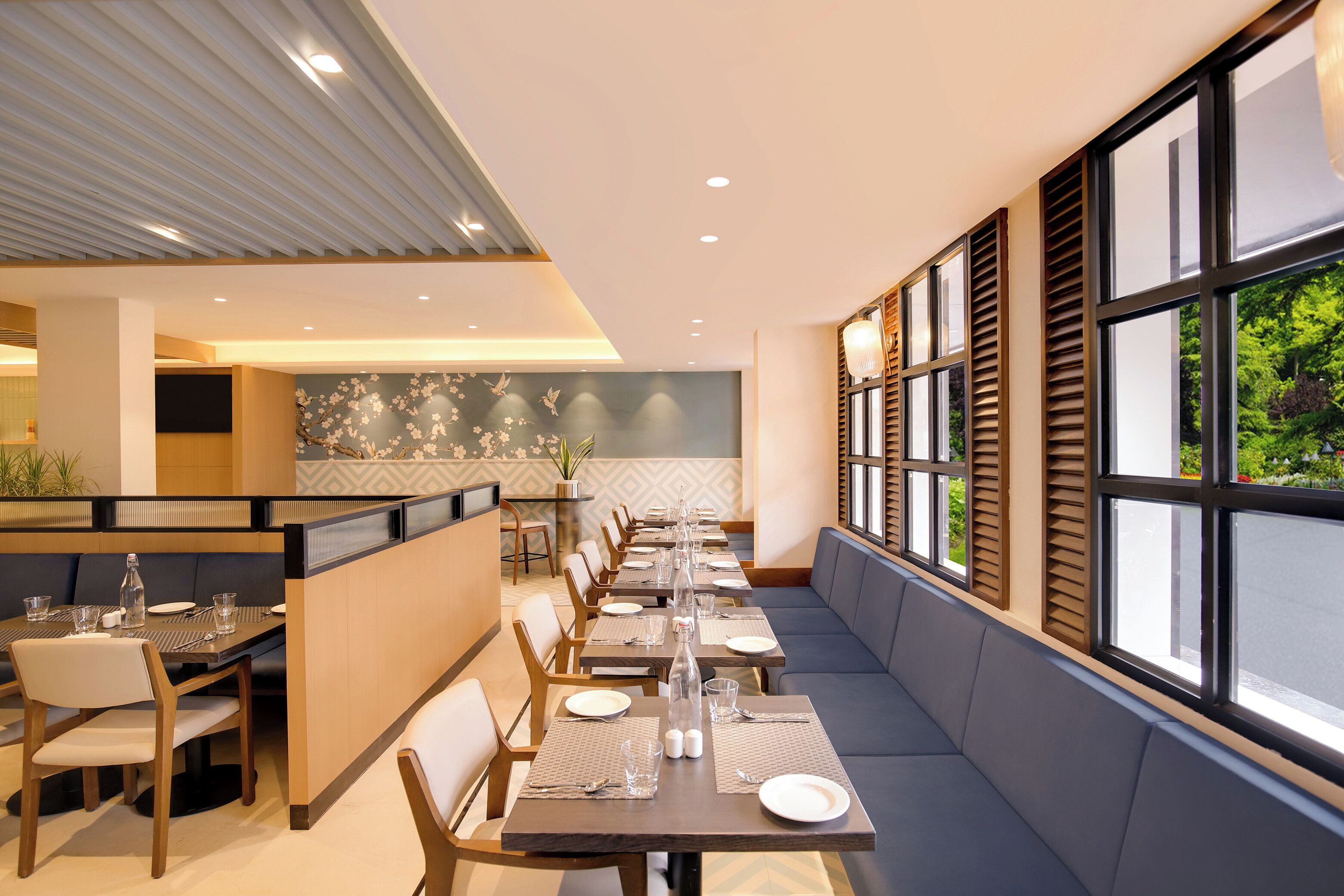 A modern dining area with long tables, cushioned seating, and large windows offering a view of greenery.
