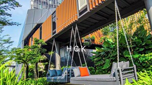 A modern outdoor seating area with swings and a cushioned bench, surrounded by lush greenery and a building with a unique architectural design. - KL City View at Trion 2 by HCK (Kuala Lumpur)