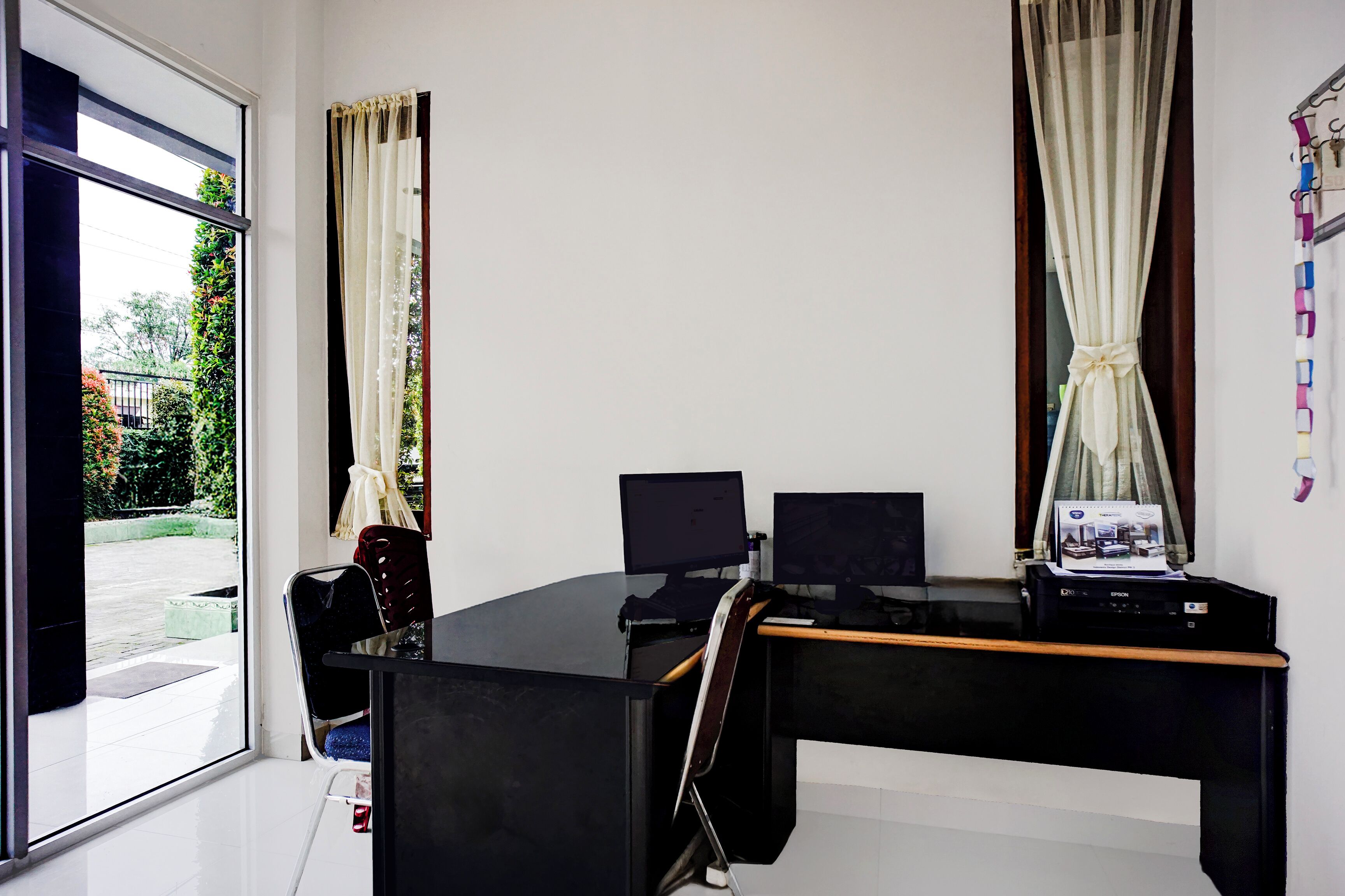 A modern office with a desk, two monitors, and a printer. There is a chair and a window with curtains.