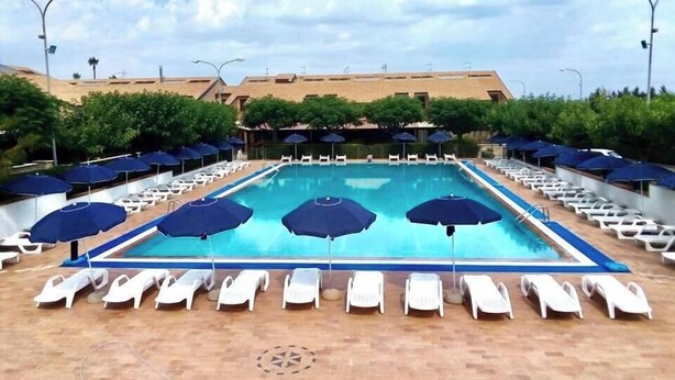Un espace piscine avec des chaises longues et des parasols, entouré de verdure et avec un bâtiment en arrière-plan.