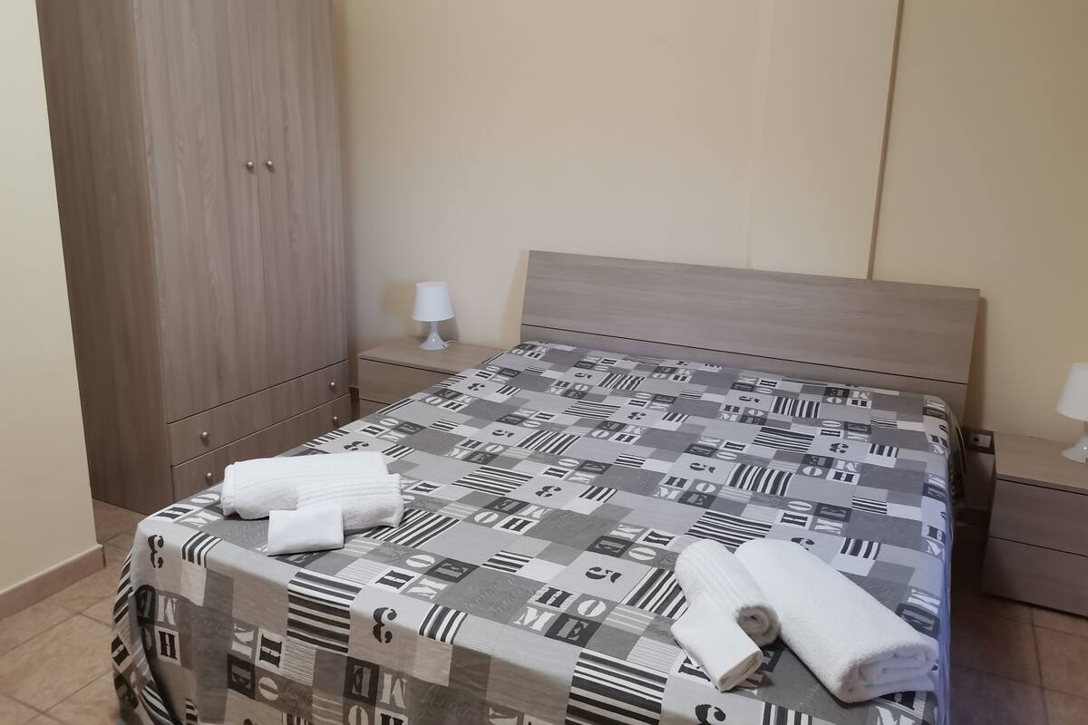 A neatly made bed with a patterned bedspread, a wooden wardrobe, and a bedside table with a lamp.