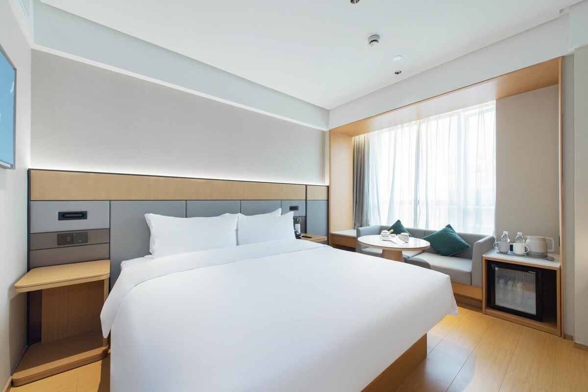 A modern hotel room with a large bed, a bedside table, a small seating area with a sofa and a table, and a minibar.