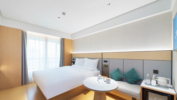 A modern hotel room with a large bed, a bedside table with a lamp, a small seating area with cushions, and a television on the wall. - Ji Hotel Guangzhou Baiyun Airport Terminal 2 (Guangzhou)