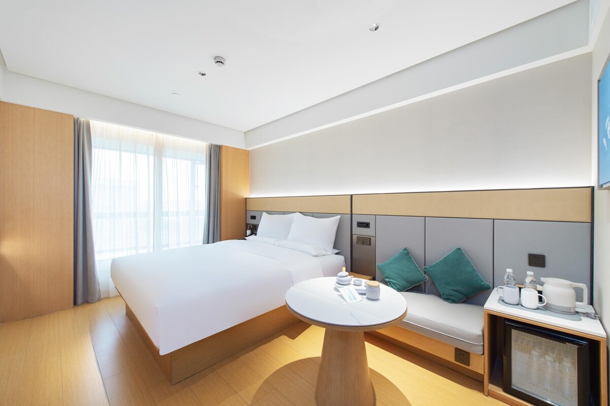 A modern hotel room with a large bed, a bedside table with a lamp, a small seating area with cushions, and a television on the wall.