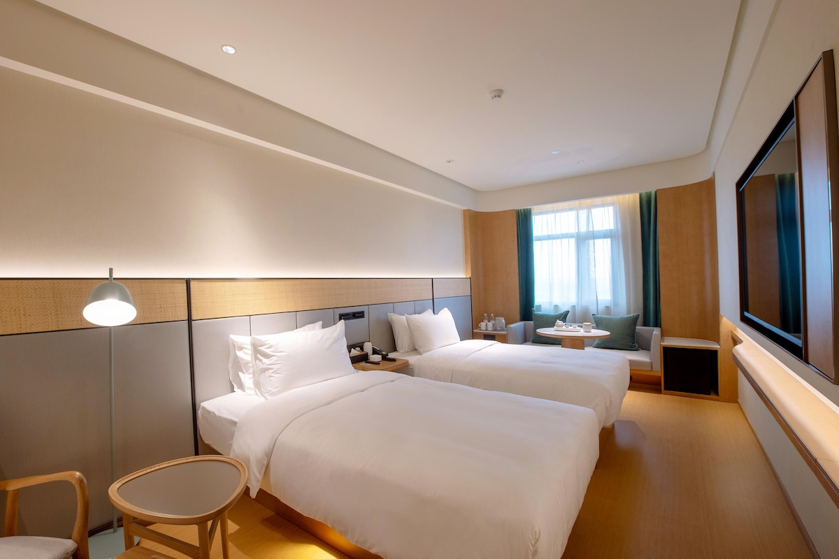 A hotel room with two beds, a desk, a chair, a TV, and a window with curtains.