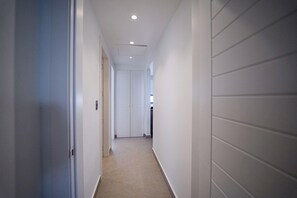 Un couloir étroit et bien éclairé, aux murs blancs et au sol carrelé, qui mène à une porte au fond.