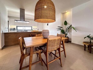 Une cuisine moderne avec des armoires blanches, un four à micro-ondes et une cuisinière. On y trouve un coin repas avec une table et des chaises en bois, et un grand luminaire suspendu en fibres tressées au-dessus de la table.