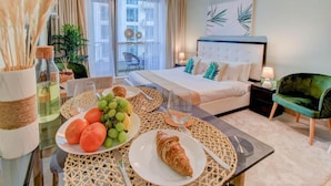 Ein Hotelzimmer mit einem Bett, einem Esstisch mit Früchten und einem Croissant, einem grünen Sessel und einem Balkon mit Blick.