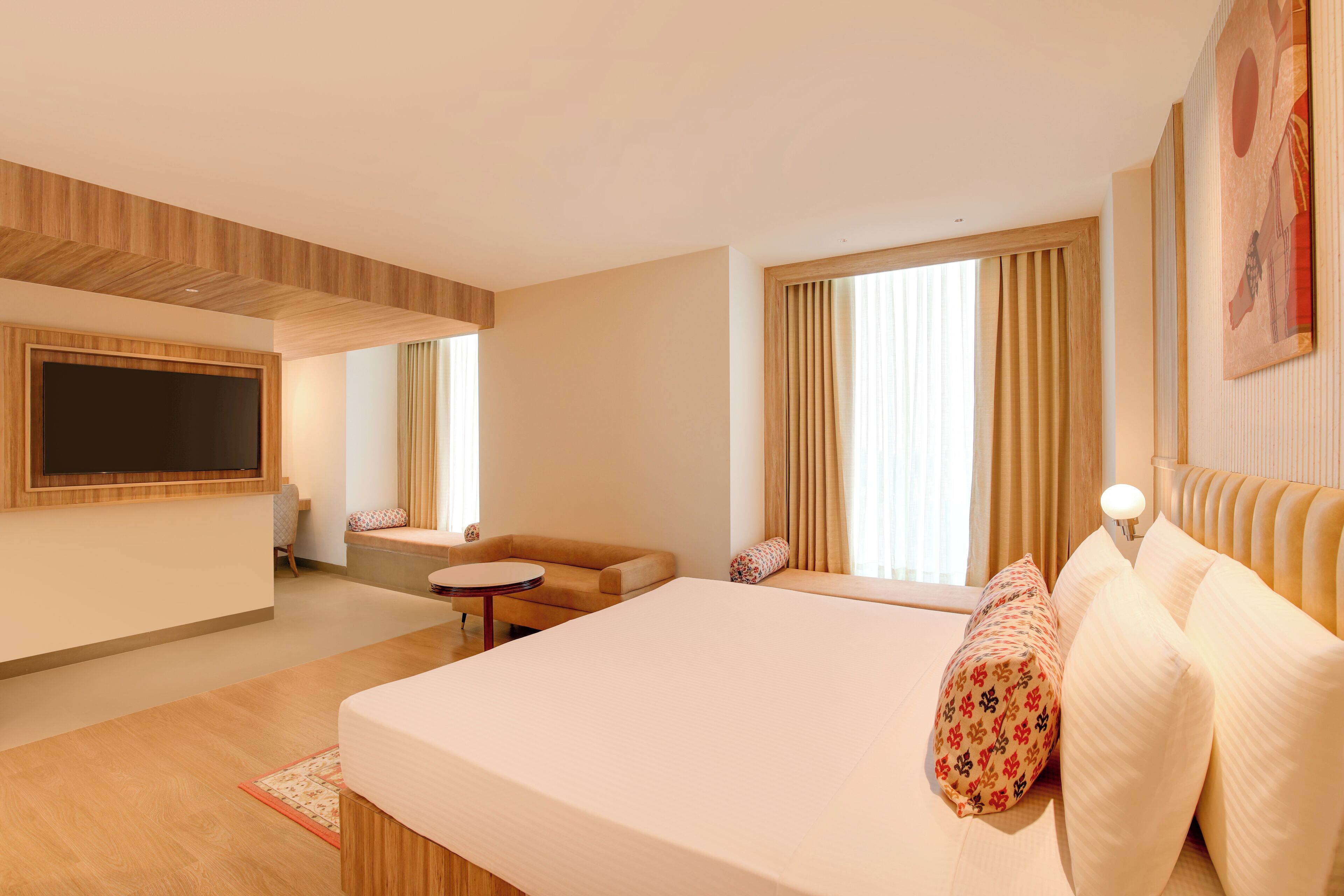 A modern hotel room with a large bed, a sofa, a small table, a TV mounted on the wall, and large windows with curtains.