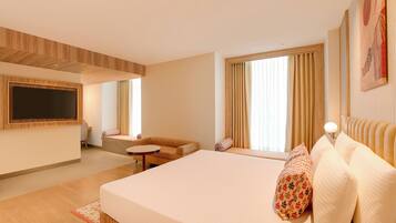 A modern hotel room with a large bed, a sofa, a small table, a TV mounted on the wall, and large windows with curtains.