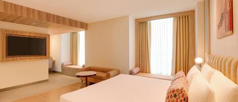 Habitación de hotel moderna con una cama grande, un sofá, una mesita, un televisor montado en la pared y ventanas amplias con cortinas.