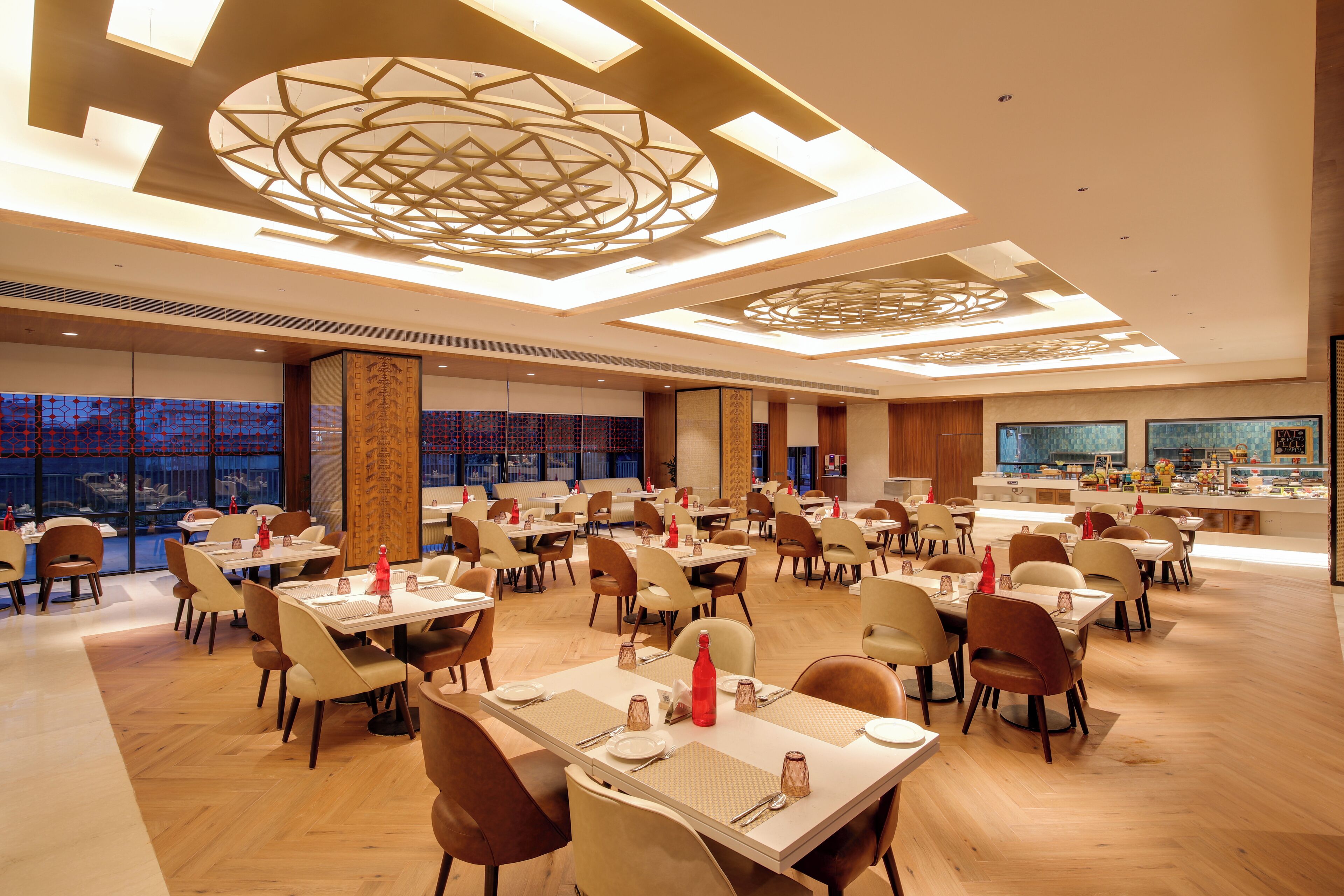 Een ruim restaurant met meerdere tafels en stoelen, een modern plafondontwerp en uitzicht op de stad door grote ramen.