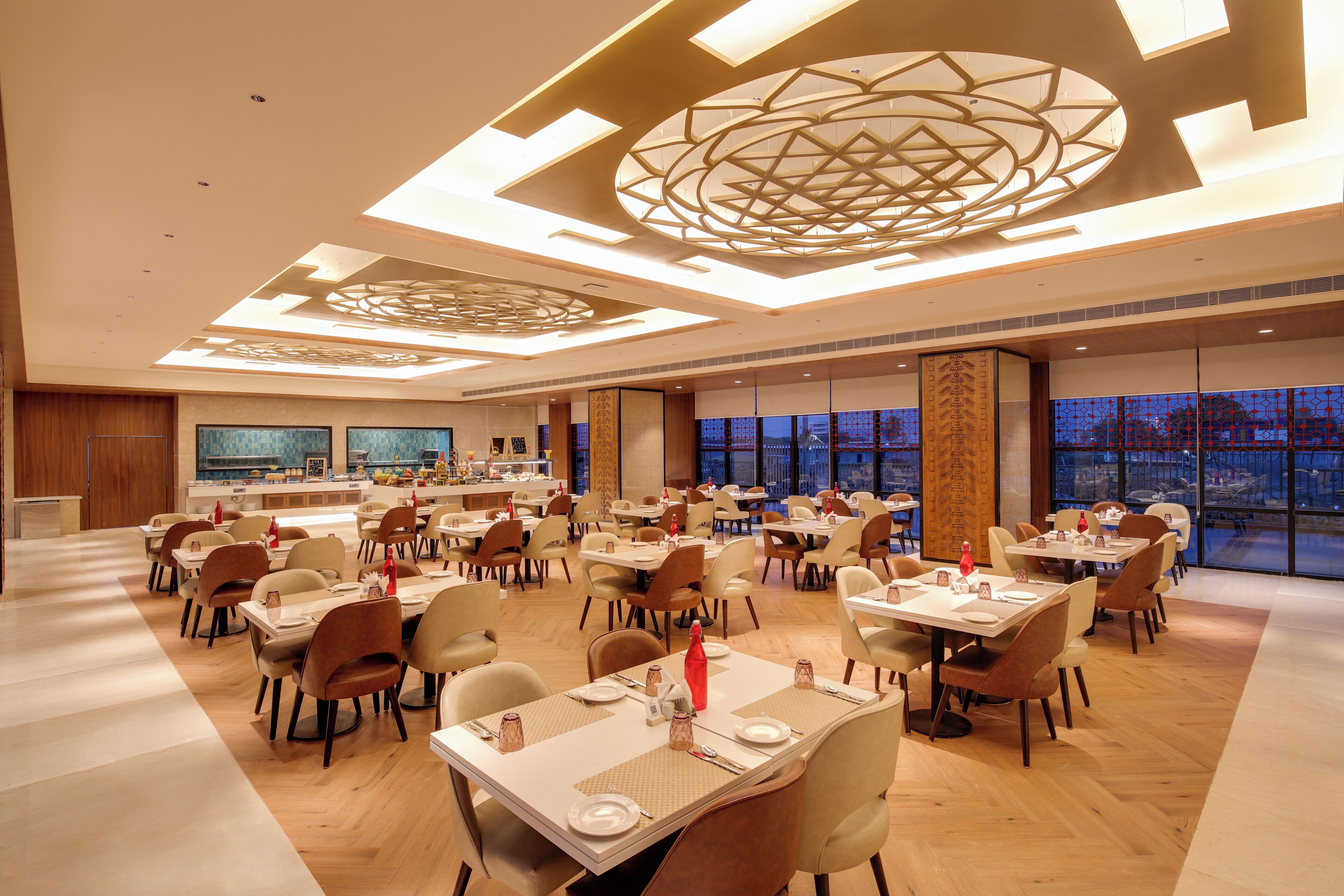A spacious dining area with multiple tables and chairs, a central decorative ceiling feature, and large windows offering a view of the cityscape.