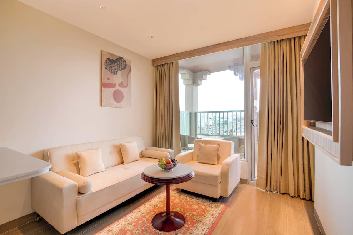 A modern hotel room with a sofa, a small round table with a fruit bowl, and a balcony with curtains.